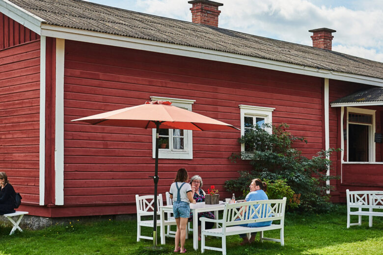 Hinttalan kotiseutumuseon punainen kahvilarakennus, jonka edessä ihmisiä kahvilla valkoisilla puutarhakalusteilla.