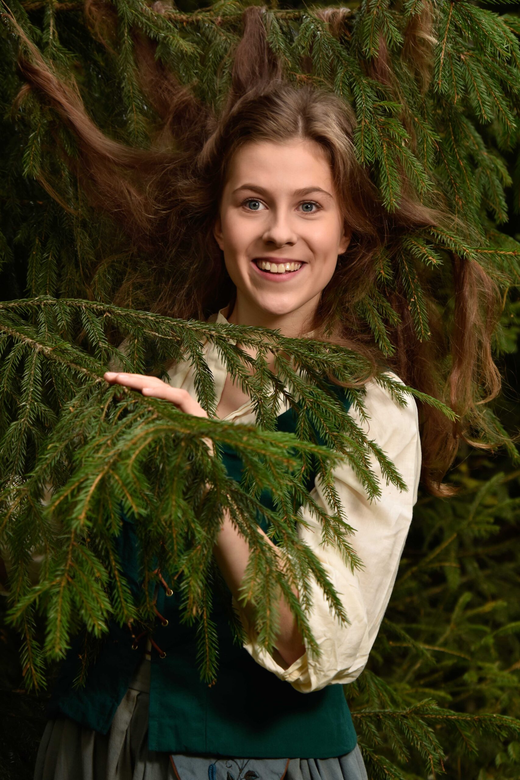 Nuori nainen katsoo kuusen oksan takaa metsässä ja hymyilee.
